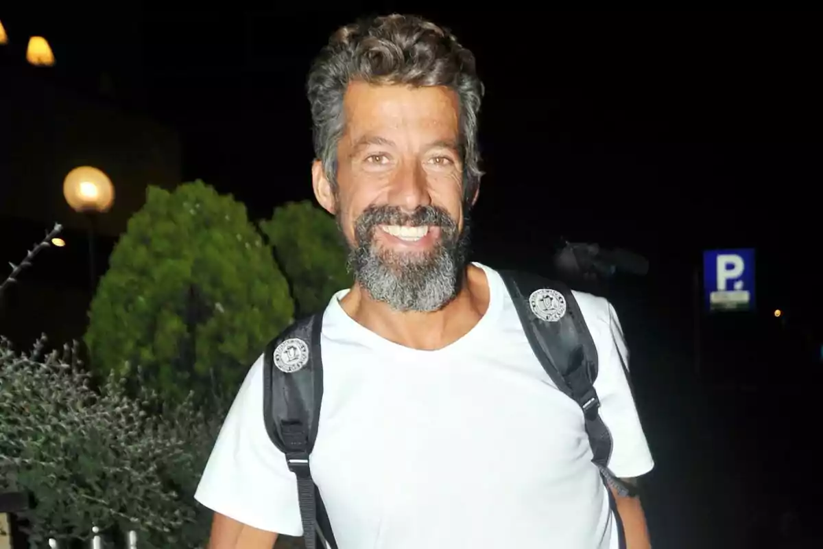 Hombre con barba y cabello rizado, sonriendo, vestido con camiseta blanca y mochila negra, en un entorno nocturno con vegetación y luces de fondo.