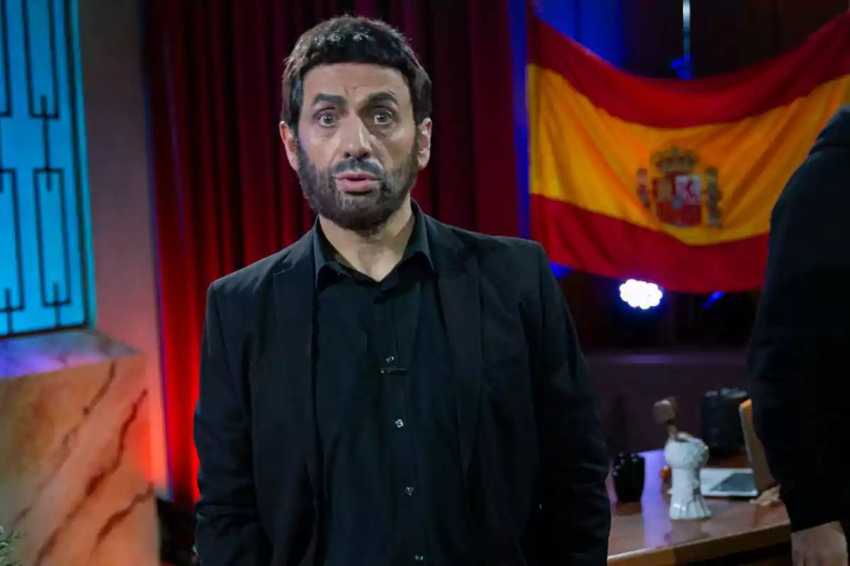 José Mota con barba y traje oscuro está de pie frente a una bandera de España en un escenario iluminado.