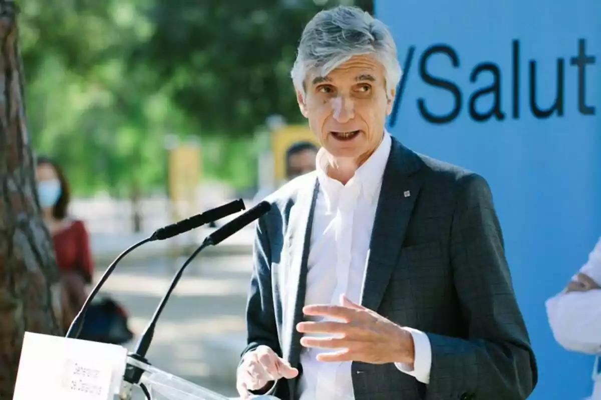 A gray-haired man speaking at an outdoor podium with a background of trees and a blue sign. A gray-haired man speaking at an outdoor podium with a background of trees and a blue sign.