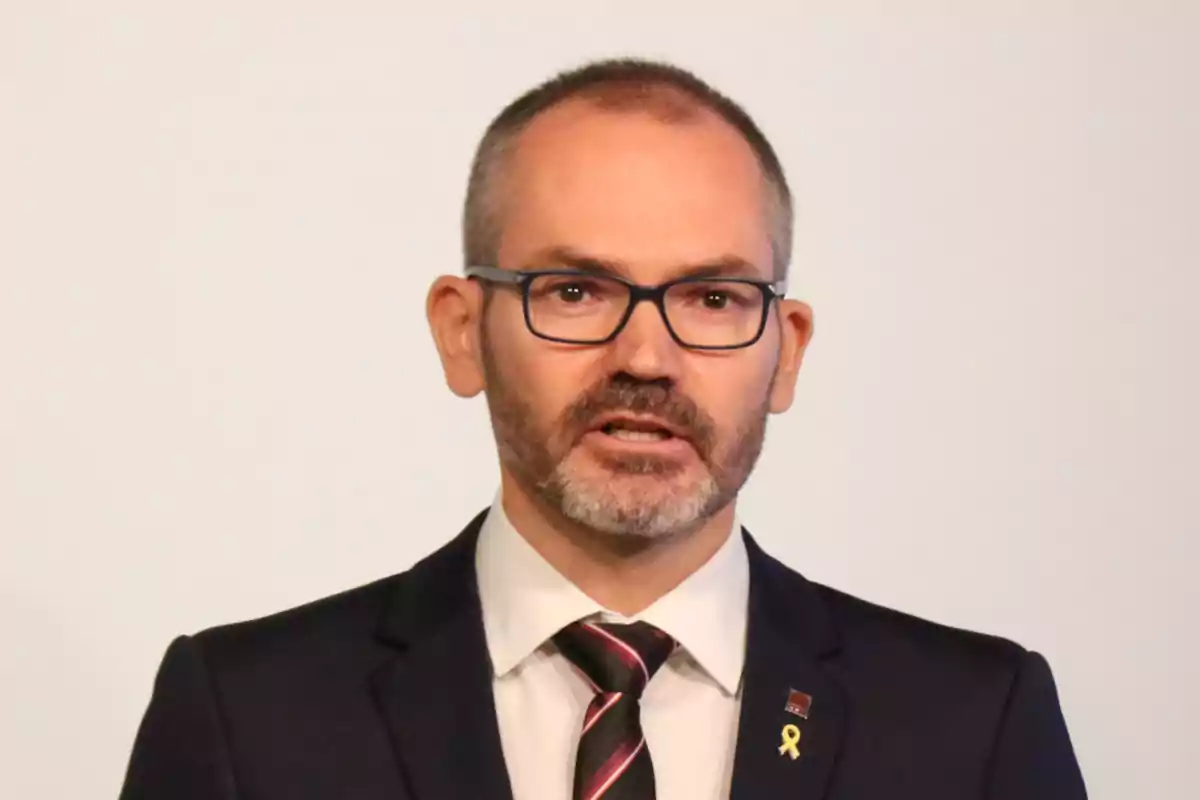 Un hombre con gafas y barba hablando mientras viste un traje oscuro y corbata.