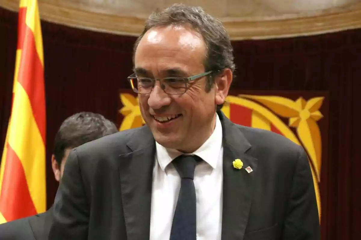 Josep Rull amb ulleres i vestit fosc somrient davant d'una bandera catalana.