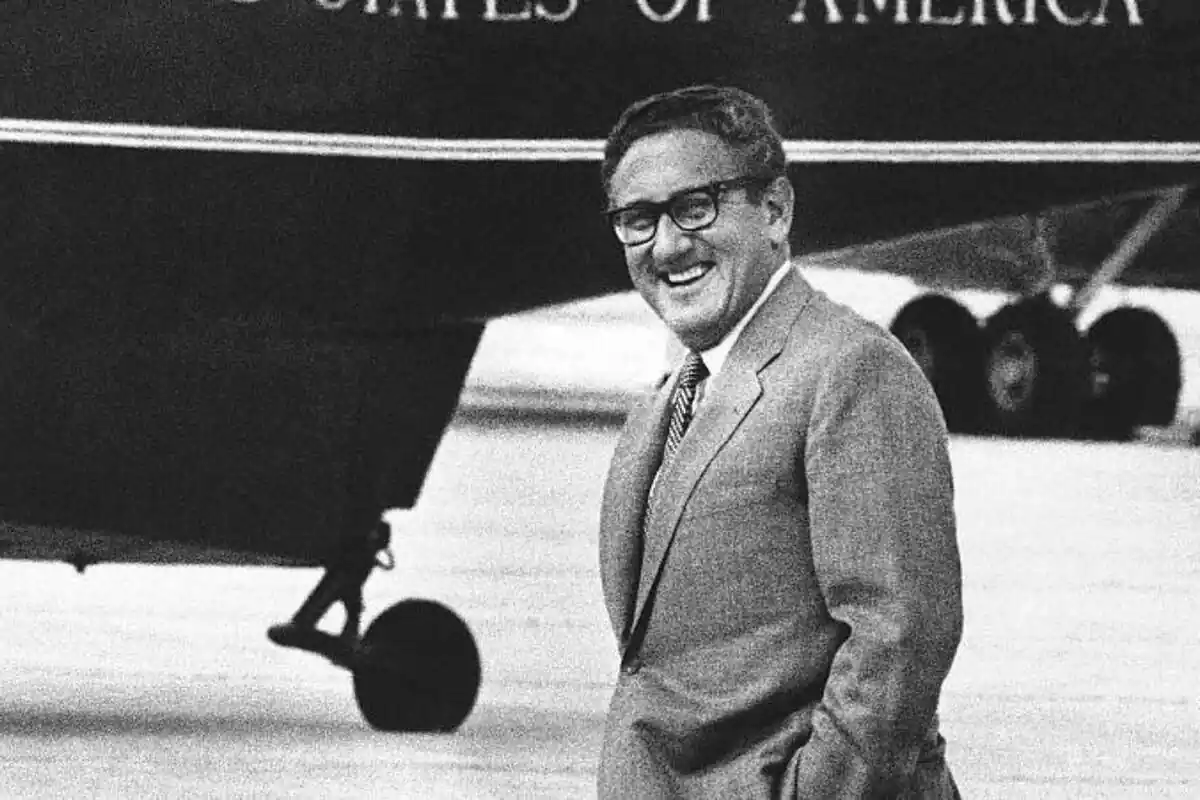 Henry Kissinger somrient a la càmera amb un avió dels EUA darrere