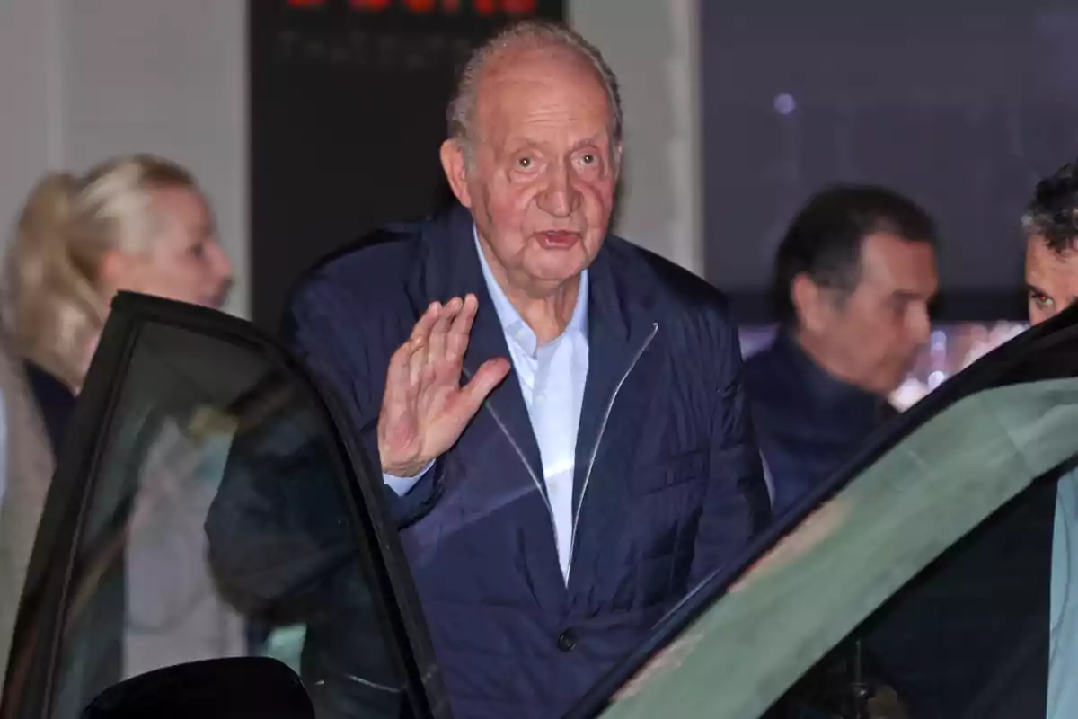 Joan Carles I amb jaqueta blava saluda mentre es troba al costat d'un automòbil, amb altres persones al fons. Joan Carles I amb jaqueta blava saluda mentre es troba al costat d'un automòbil, amb altres persones al fons.