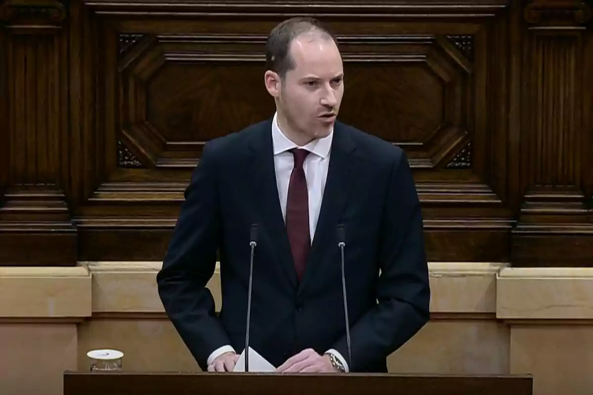 Un home de vestit fosc i corbata vermella parla des d'un faristol de fusta amb micròfons davant seu. Un home de vestit fosc i corbata vermella parla des d'un faristol de fusta amb micròfons davant seu.