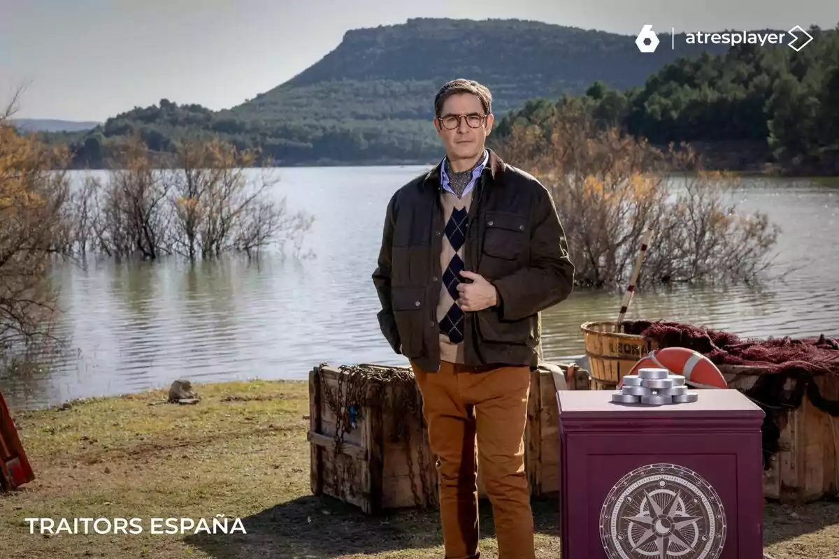 Juanra Bonet dreta al costat d'un llac amb turons i arbres al fons, envoltat de caixes de fusta i una taula amb un símbol a Traitors España.