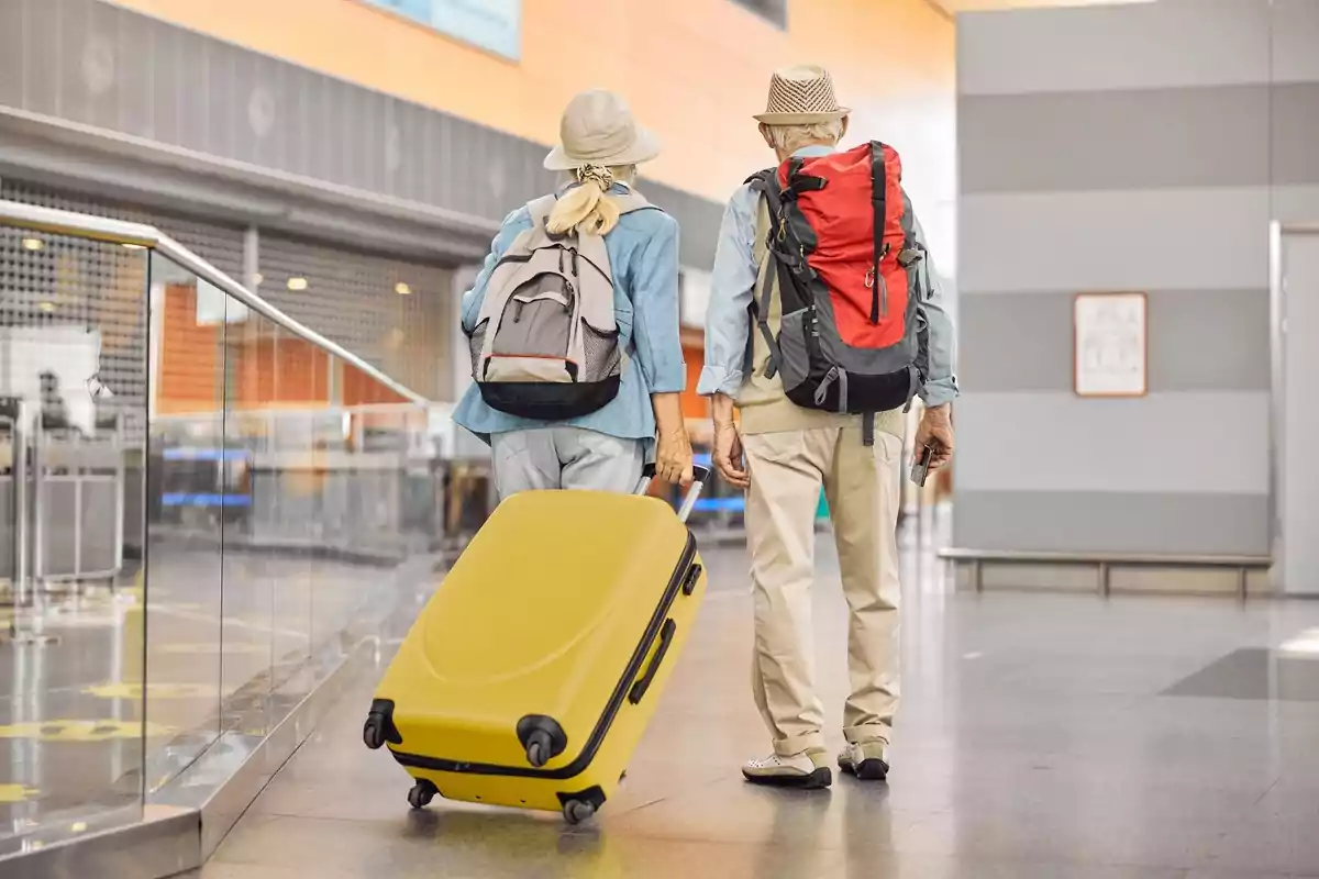 Dues persones grans amb motxilles i una maleta groga caminant per un aeroport. Dues persones grans amb motxilles i una maleta groga caminant per un aeroport.