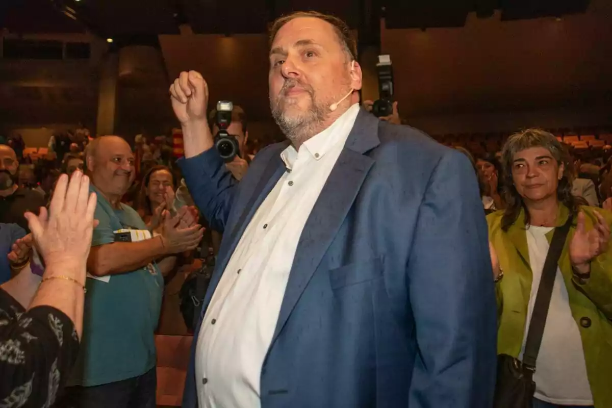 Oriol Junqueras amb vestit blau i camisa blanca aixeca el braç mentre és aplaudit per una multitud en un esdeveniment, amb fotògrafs capturant el moment.