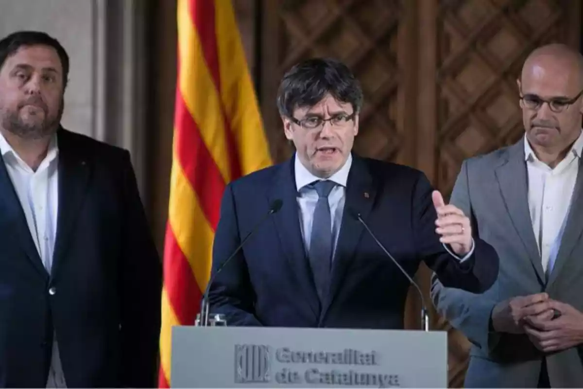 pla mig de Puigdemont parlant davant de dos micròfons, darrere seu hi ha Junqueras i Romeva seriosos escoltant-lo