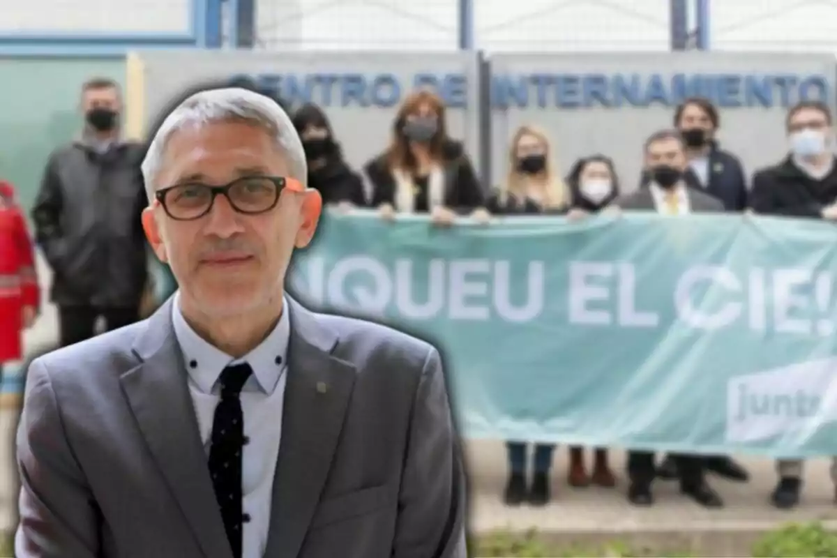 Un home amb ulleres i vestit està en primer pla, mentre que al fons hi ha un grup de persones amb màscares sostenint una pancarta davant d'un edifici.