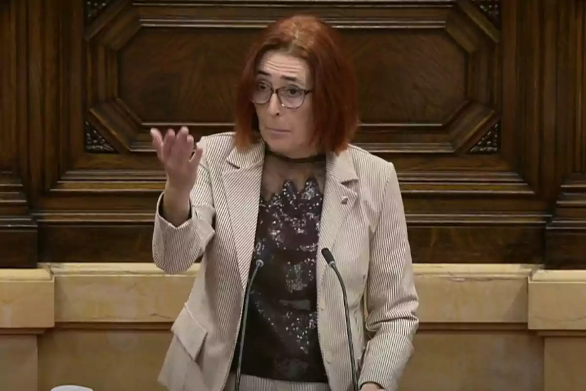 Una persona con cabello rojo y gafas está hablando en un podio de madera, gesticulando con la mano.