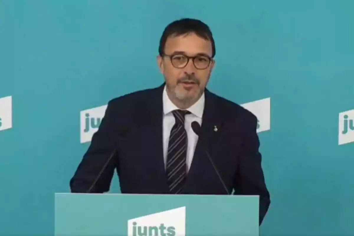 A man with glasses and a dark suit speaks at a podium with a blue background featuring the "junts" logo.