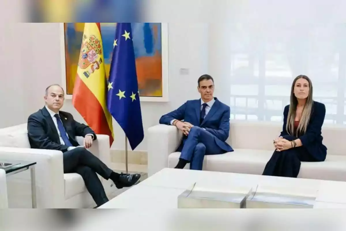 Tres persones amb vestimenta formal assegudes en sofàs blancs en una sala de reunions amb les banderes d’Espanya i de la Unió Europea al fons Tres persones amb vestimenta formal assegudes en sofàs blancs en una sala de reunions amb les banderes d’Espanya i de la Unió Europea al fons
