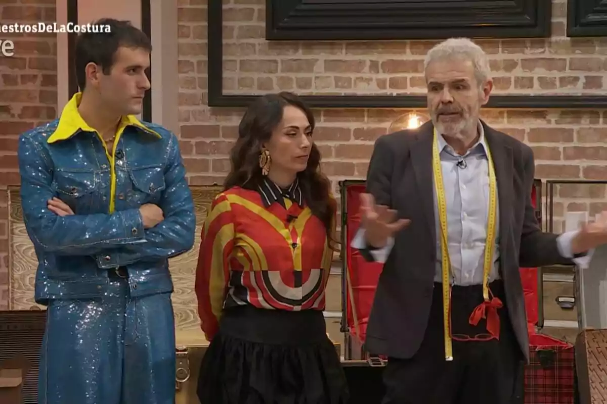 Palomo, María Escoté y Lorenzo Caprile de pie en un set de televisión con fondo de ladrillos, una de ellas lleva un traje azul brillante, otra una blusa colorida y la tercera un traje con una cinta métrica alrededor del cuello.