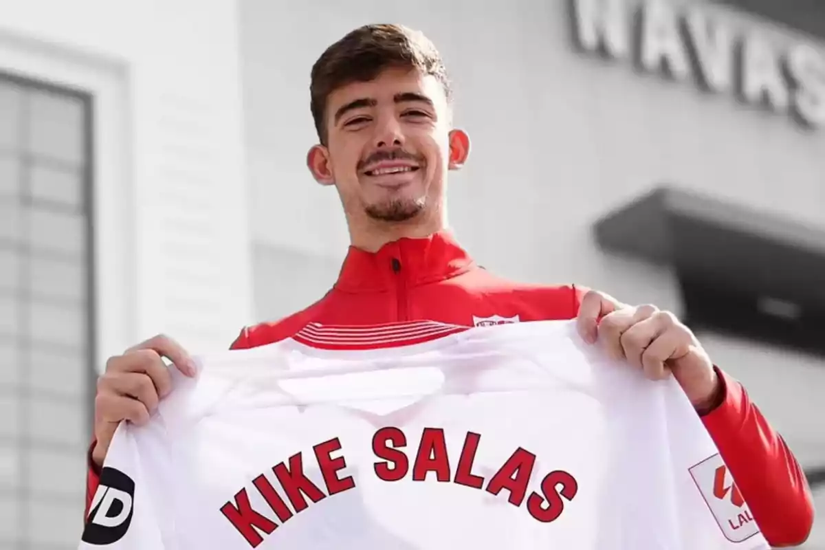 Un hombre sonriente sostiene una camiseta blanca con el nombre "Kike Salas" en letras rojas.