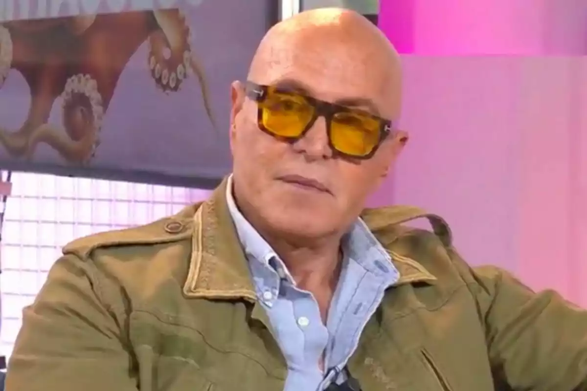 Kiko Matamoros calb amb ulleres de sol grogues i jaqueta verda està assegut en un entorn il·luminat amb llums rosades i un cartell de fons que mostra el programa 'Tentacles'.