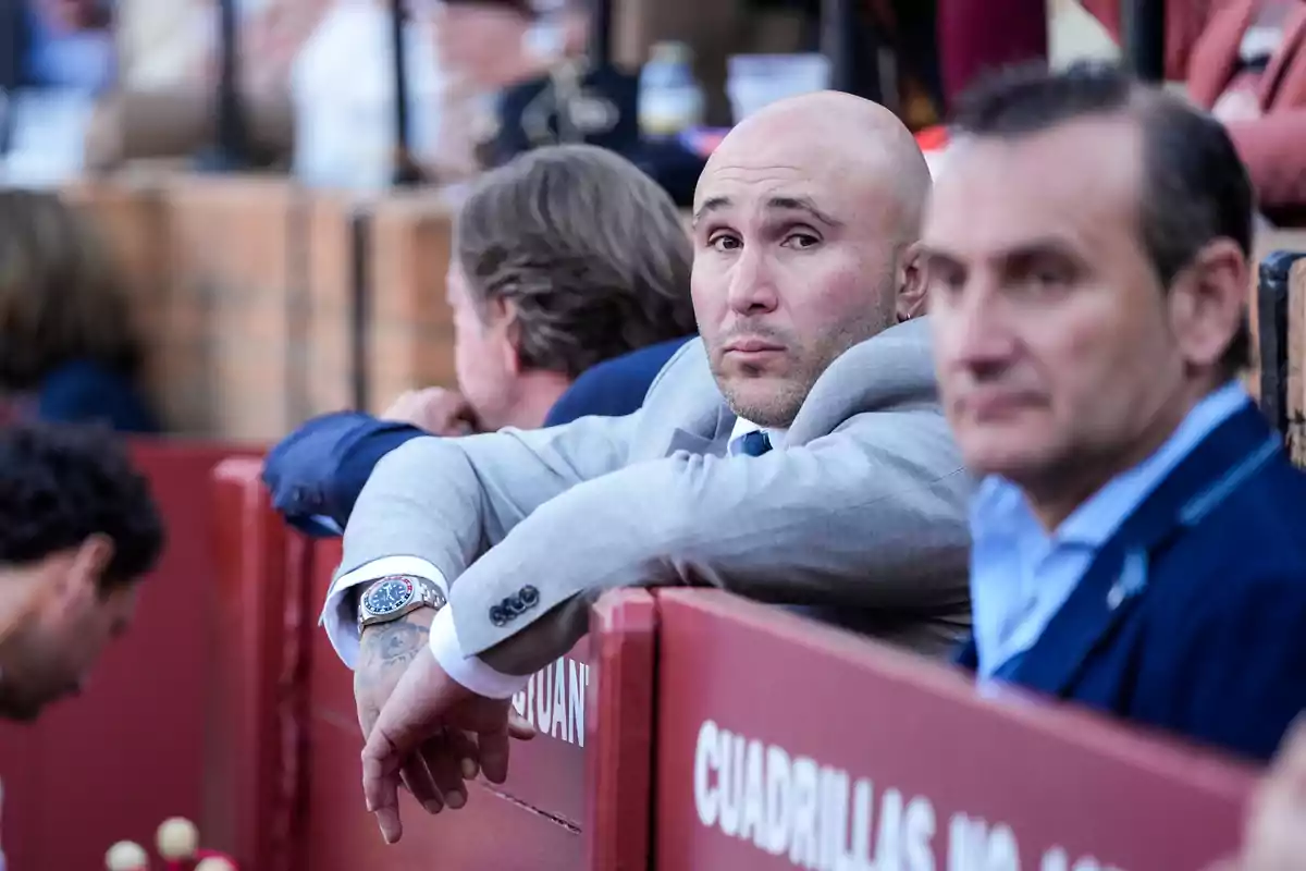 A man in a gray suit with a watch on his wrist watches from a barrier at a public event. A man in a gray suit with a watch on his wrist watches from a barrier at a public event.
