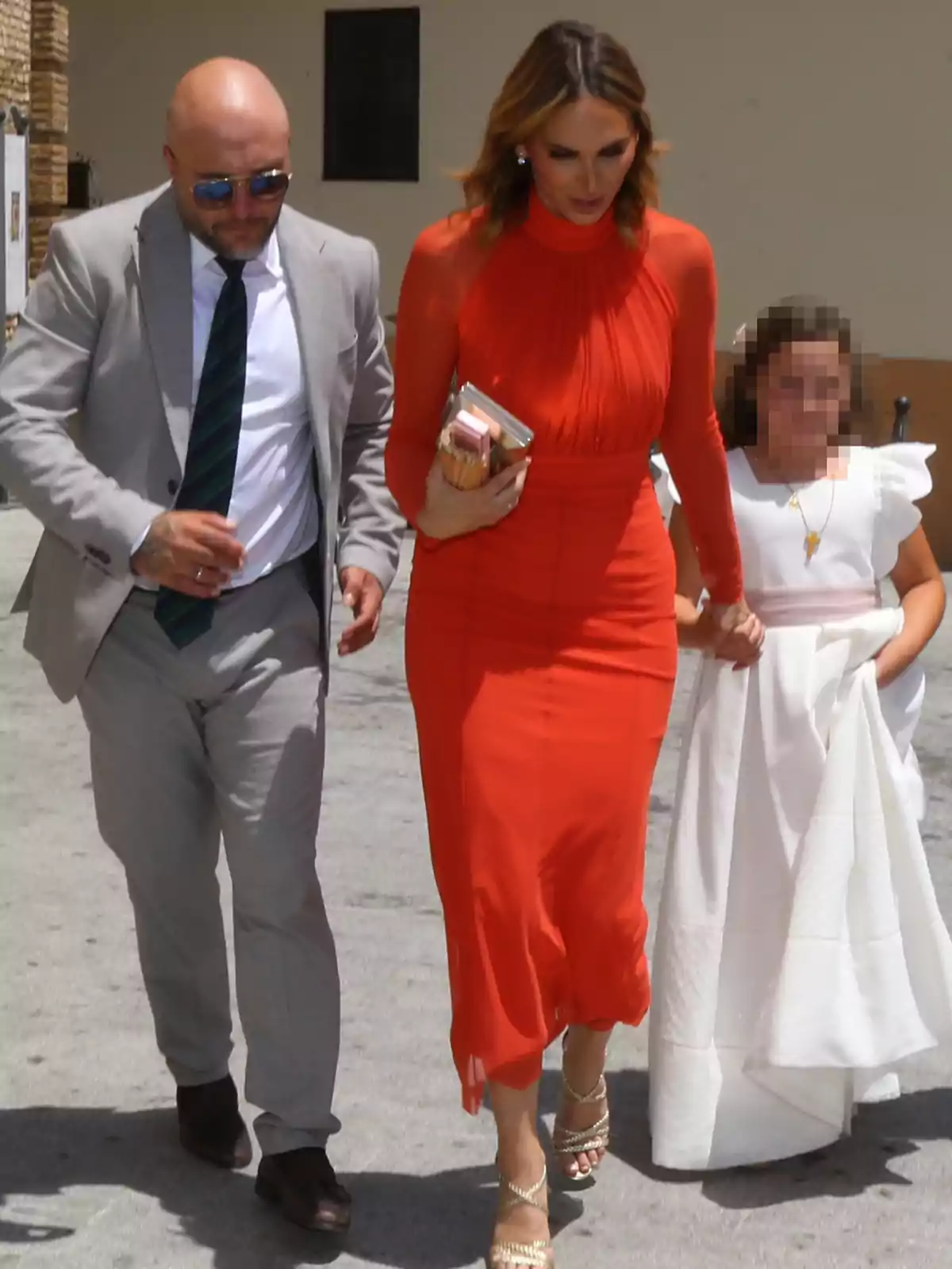Kiko Rivera in a gray suit and tie, Irene Rosales in a red dress and gold sandals, and a girl dressed in white walk together outdoors. Kiko Rivera in a gray suit and tie, Irene Rosales in a red dress and gold sandals, and a girl dressed in white walk together outdoors.