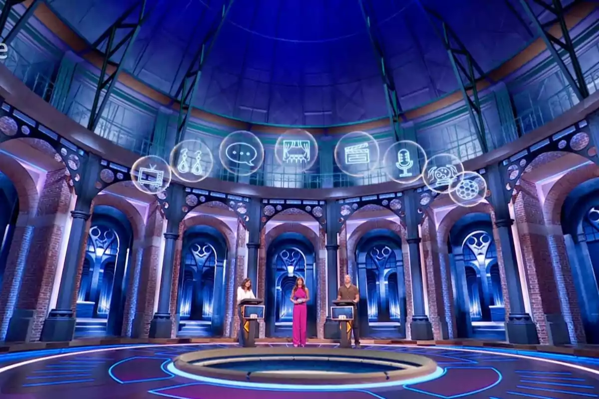 Escenari futurista de 'La Connexió' amb tres persones al centre, envoltades de columnes i arcs, i diversos icones flotant sobre elles en un ambient il·luminat amb llums blaves i morades. Escenari futurista de 'La Connexió' amb tres persones al centre, envoltades de columnes i arcs, i diversos icones flotant sobre elles en un ambient il·luminat amb llums blaves i morades.