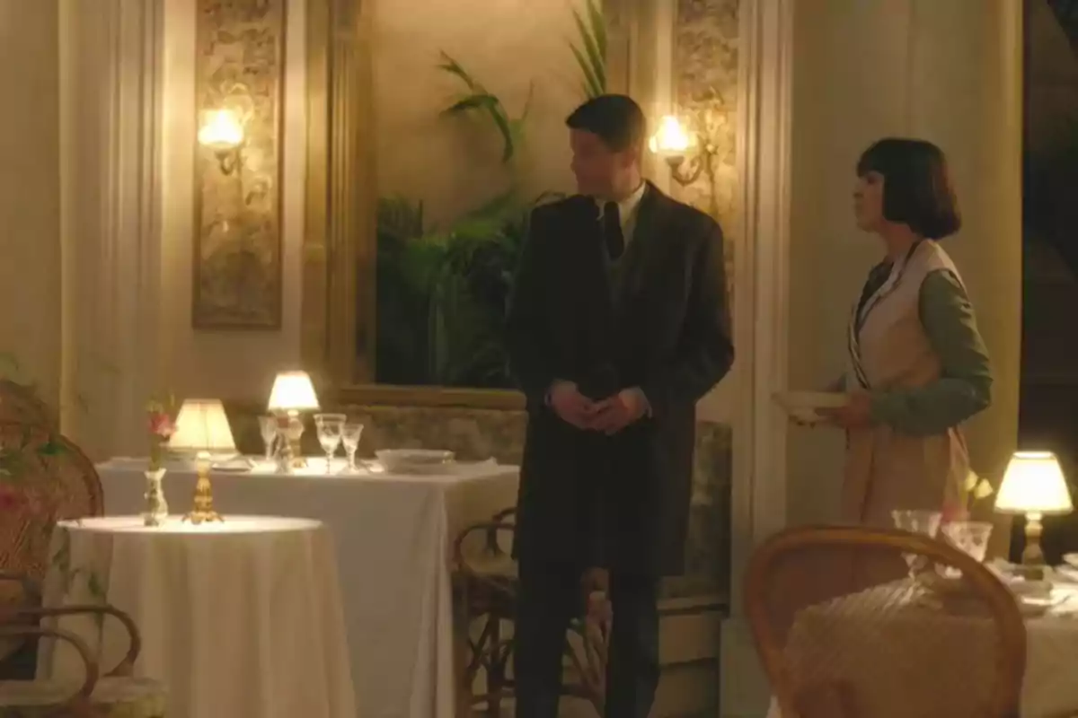 Juliol i Elena estan drets en un elegant restaurant amb taules decorades amb estovalles blanques, copes i làmpades de taula, mentre conversen en un ambient càlid i acollidor. Captura de 'La Favorita 1922'. Juliol i Elena estan drets en un elegant restaurant amb taules decorades amb estovalles blanques, copes i làmpades de taula, mentre conversen en un ambient càlid i acollidor. Captura de 'La Favorita 1922'.