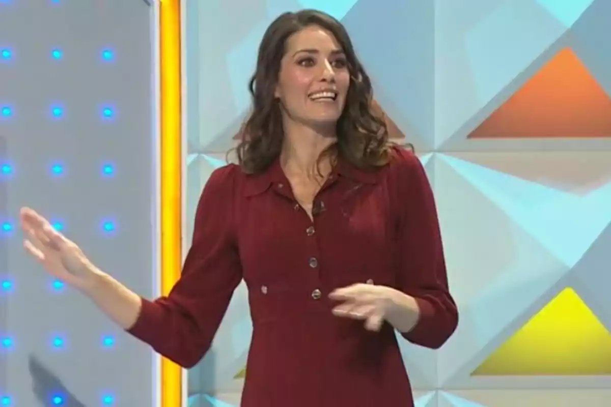 Laura Moure amb vestit vermell somriu mentre gesticula en un plató de televisió amb fons geomètric i llums blaves. Captura de 'La Ruleta de la Suerte'