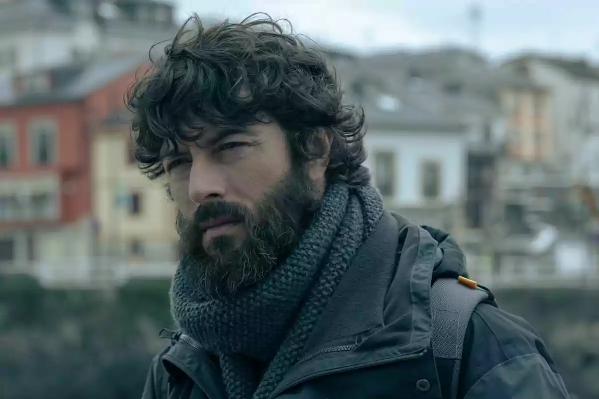 Javier Rey con barba y cabello rizado, usando una bufanda gris y una chaqueta, con un fondo de edificios desenfocados en La última noche en Tremor.