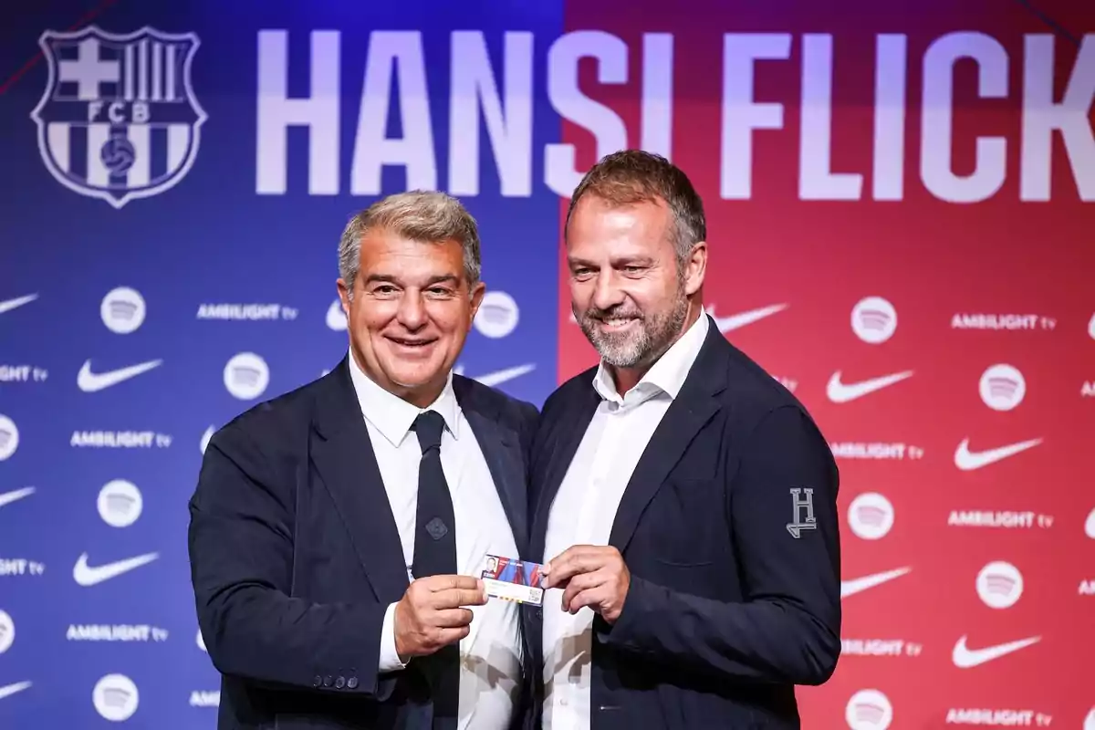 Two men in dark suits pose together holding a card, with a background showing logos of Nike, Spotify and Ambilight TV, and the FCB crest.