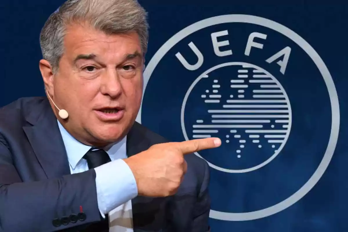Home de vestit i corbata, amb un micròfon de diadema, assenyalant amb el dit índex cap a un logotip de la UEFA al fons.