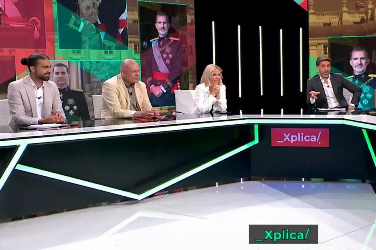 Un grup de persones encapçalades per José Yélamo assegudes en un plató de televisió amb pantalles de fons mostrant imatges d'una figura en uniforme militar i el logotip del programa _Xplíca! a l'escriptori.
