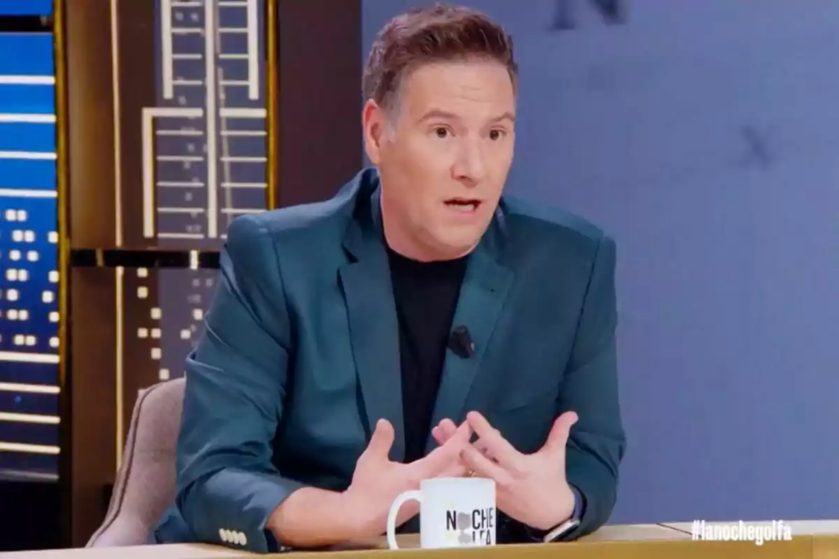 Carlos Latre amb jaqueta blau fosc està assegut en un plató de televisió, gesticulant mentre parla i té una tassa blanca sobre la taula a 'La nit golfa'.