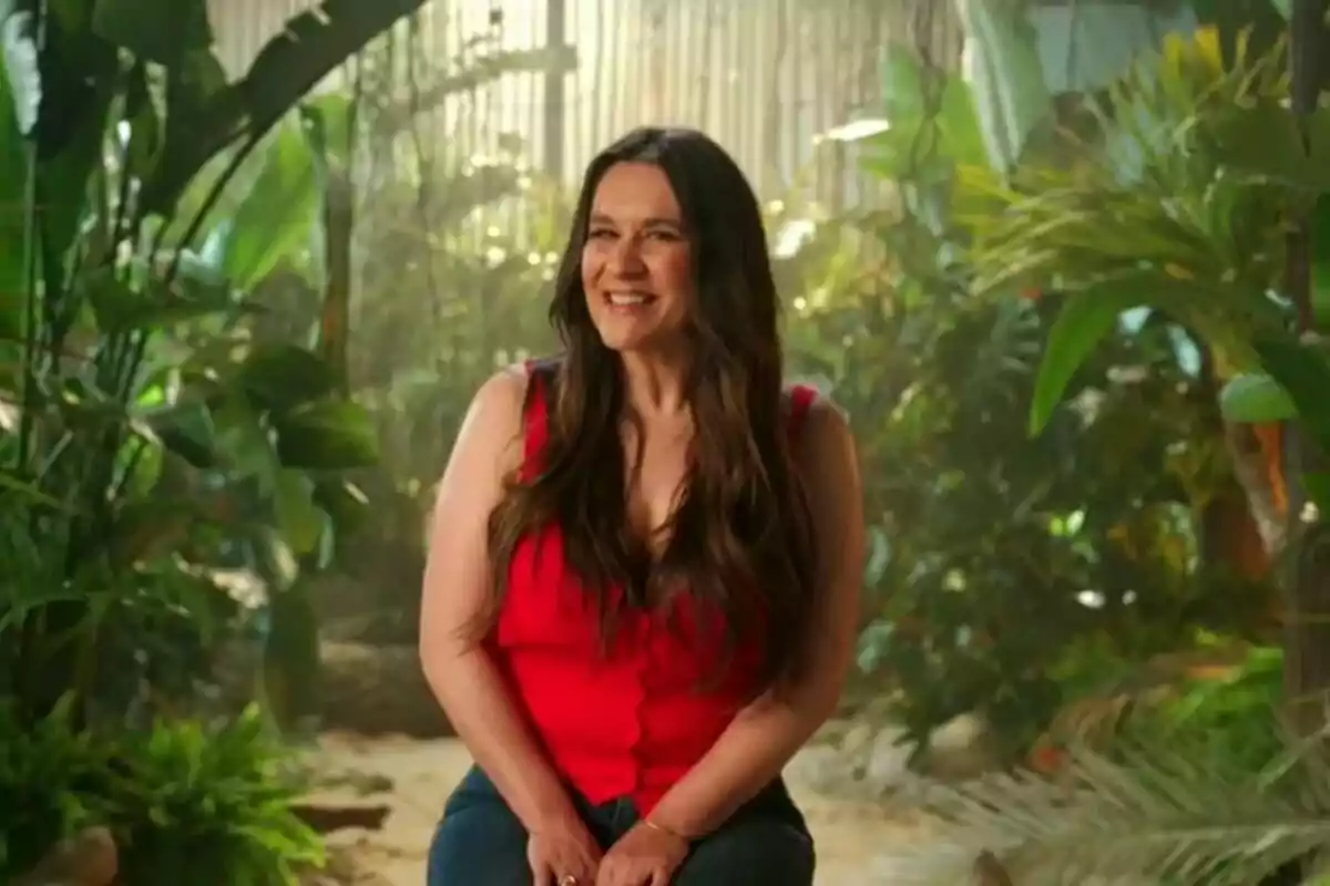 Laura Cuevas sonriente con cabello largo y oscuro, vestida con una blusa roja, sentada en un entorno lleno de plantas verdes en su confirmación de Supervivientes.