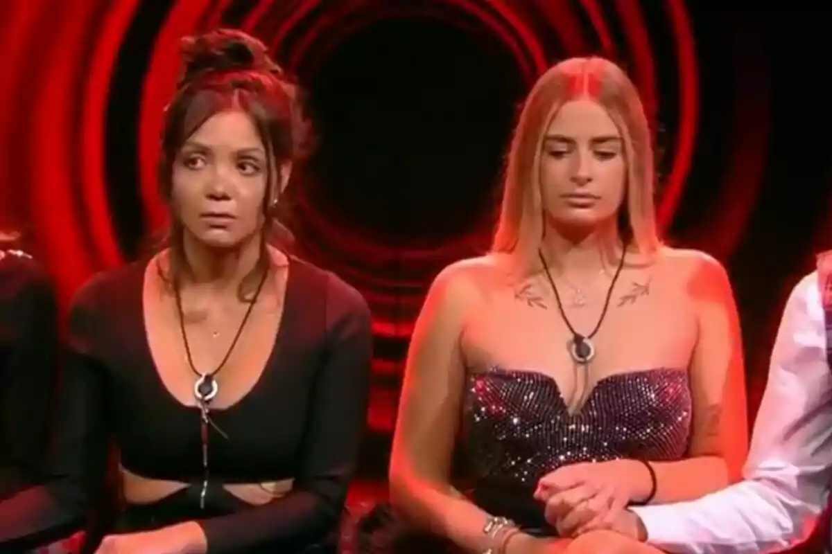 Laura Galera i Daniela assegudes a la sala d'expulsió de Gran Hermano amb llums vermelles de fons, una porta un vestit negre i l'altra un vestit brillant.