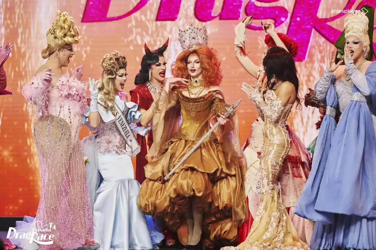 Le Cocó junto a un grupo de personas en elaborados trajes de gala celebra en un escenario con fondo brillante en Drag Race España.