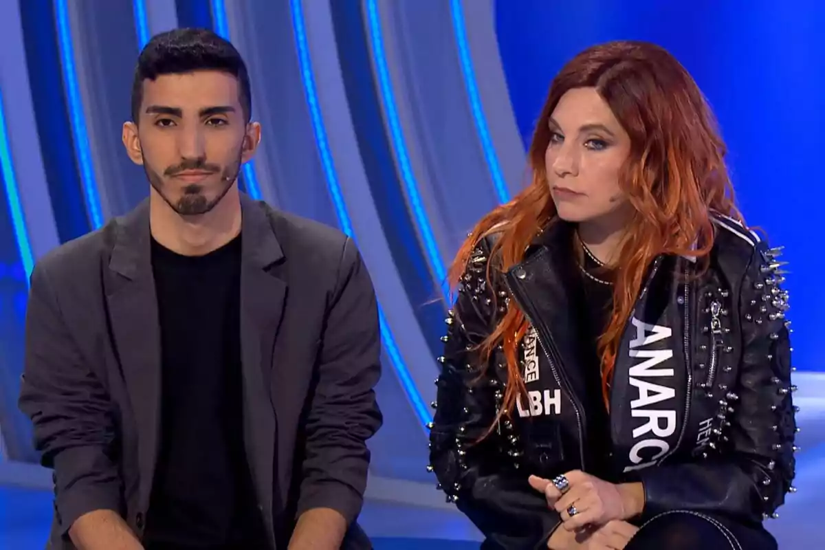 Leire Martínez i un noi asseguts a Hay en una cosa que et vull dir amb fons blau, una d'elles porta una jaqueta de cuir amb tatxes i l'altra porta un blazer fosc. Leire Martínez i un noi asseguts a Hay en una cosa que et vull dir amb fons blau, una d'elles porta una jaqueta de cuir amb tatxes i l'altra porta un blazer fosc.