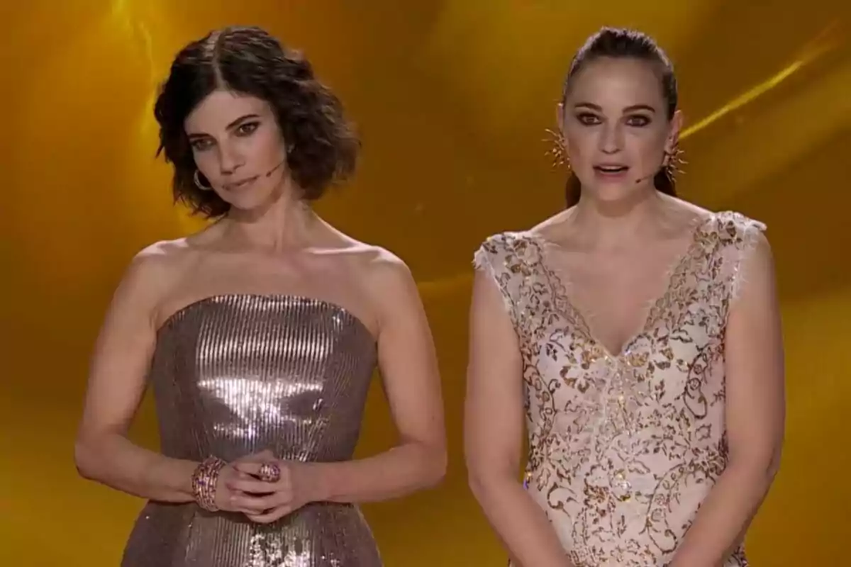 Maribel Verdú y Leonor Watling en un escenario de los Premios Goya con vestidos elegantes y micrófonos, una con un vestido plateado y la otra con un vestido blanco con detalles dorados, sobre un fondo dorado.