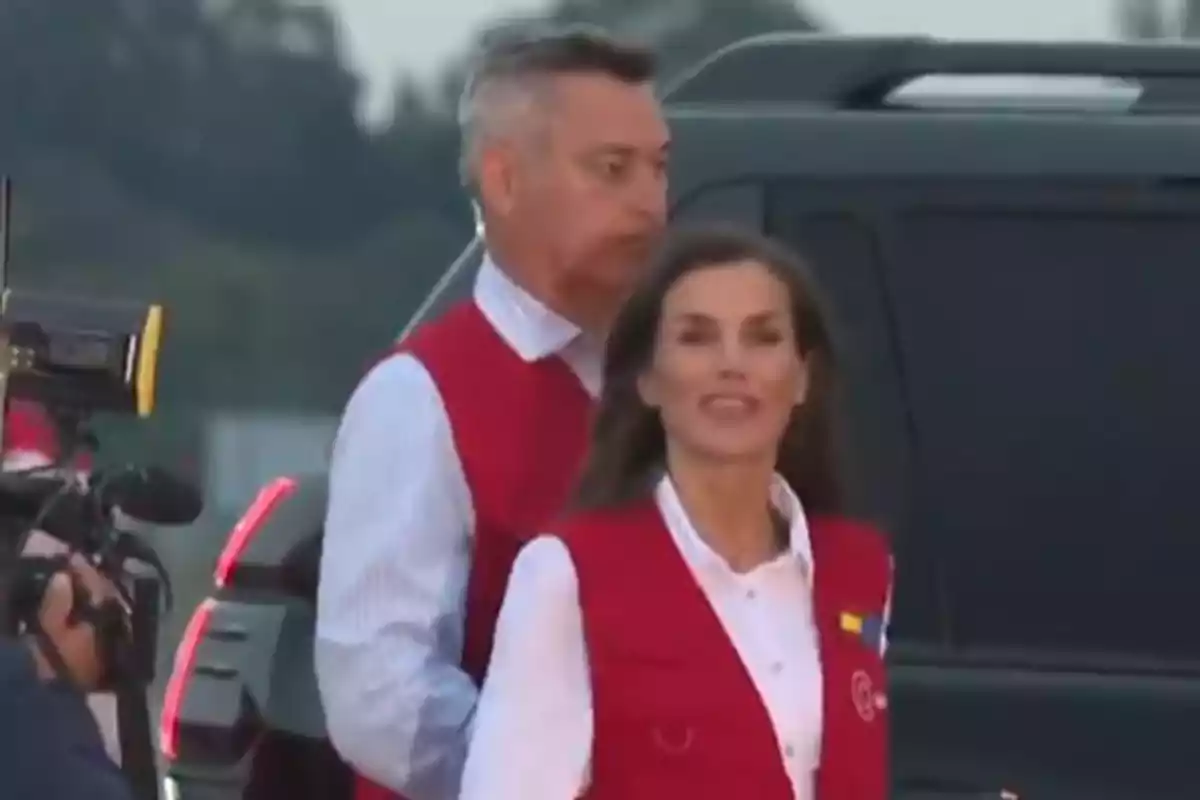Queen Letizia and a man in red vests are standing in front of a vehicle, with a camera and a microphone visible on the left. Queen Letizia and a man in red vests are standing in front of a vehicle, with a camera and a microphone visible on the left.