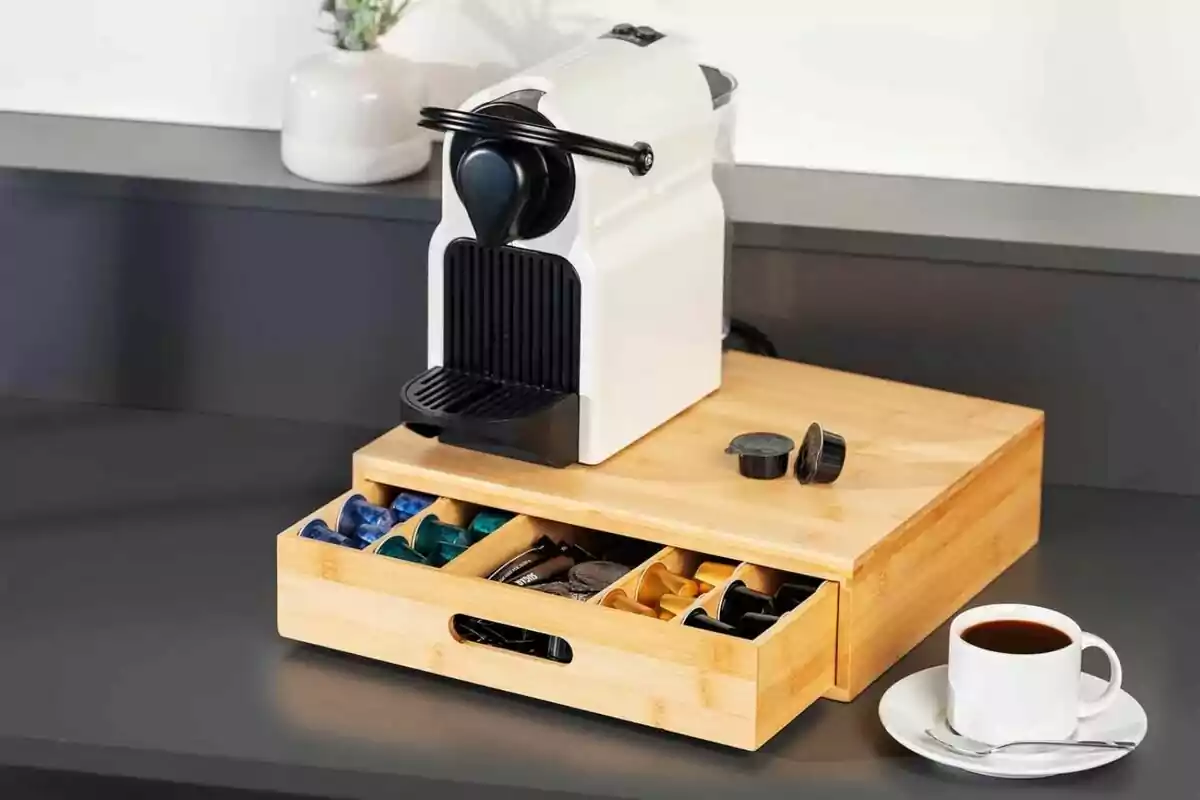 Cafetera de càpsules blanca sobre una caixa organitzadora de fusta amb càpsules de cafè i una tassa de cafè sobre una taula Cafetera de càpsules blanca sobre una caixa organitzadora de fusta amb càpsules de cafè i una tassa de cafè sobre una taula