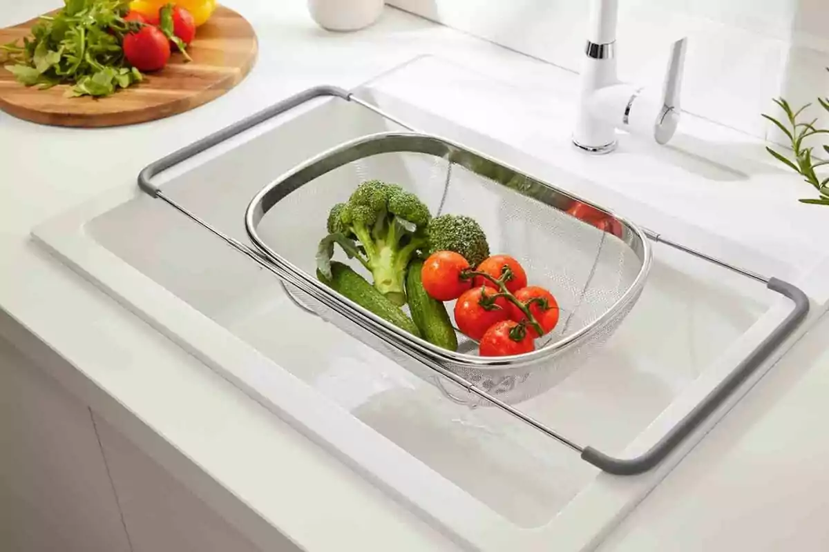 A metal strainer over a sink with broccoli, tomatoes, and cucumbers, and a cutting board with more vegetables in the background.