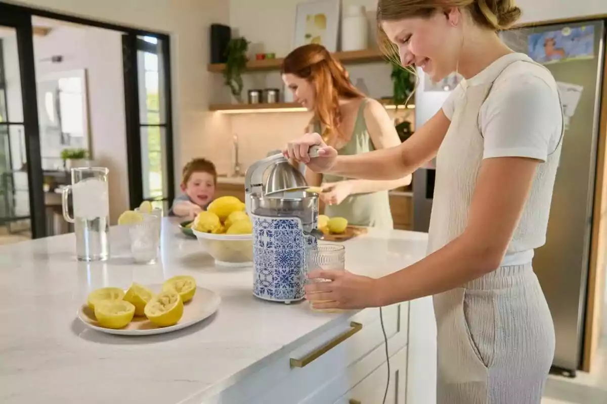 A person squeezes lemons in a modern kitchen while another person and a child are in the background, with a plate of cut lemons and a fielder of water on the countertop. A person squeezes lemons in a modern kitchen while another person and a child are in the background, with a plate of cut lemons and a fielder of water on the countertop.