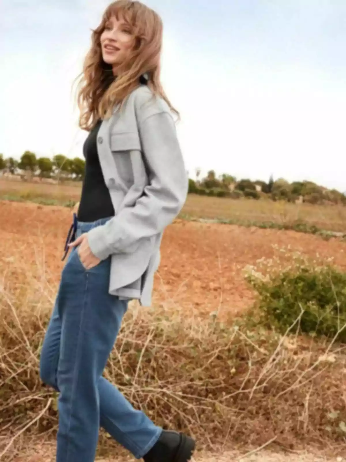 A person walking through a field in casual attire and a gray jacket. A person walking through a field in casual attire and a gray jacket.