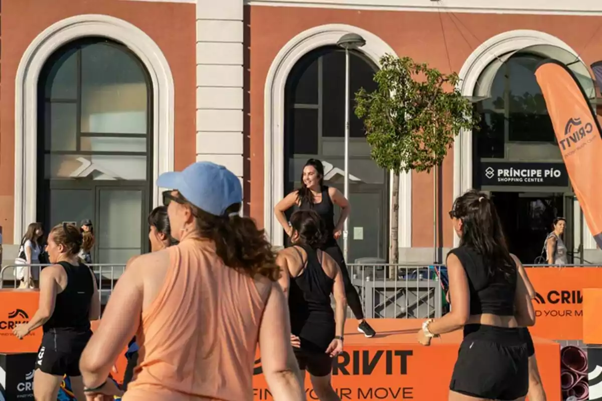 A group of women exercise outdoors in front of a building with large windows and a shopping center sign while an instructor smiles on a platform. A group of women exercise outdoors in front of a building with large windows and a shopping center sign while an instructor smiles on a platform.
