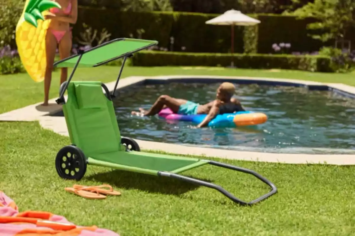 Cadira verda amb rodes i tendal a la gespa al costat d’una piscina on una persona flota en un inflable i una altra sosté un matalàs de pinya. Cadira verda amb rodes i tendal a la gespa al costat d’una piscina on una persona flota en un inflable i una altra sosté un matalàs de pinya.