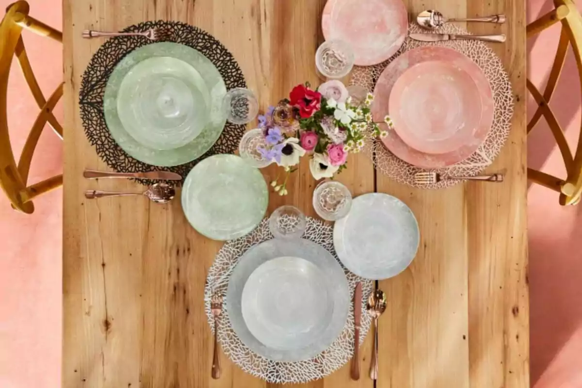 Una taula de fusta amb tres llocs disposats amb plats de colors pastís, coberts de metall i copes de vidre, decorada amb un centre de taula floral. Una taula de fusta amb tres llocs disposats amb plats de colors pastís, coberts de metall i copes de vidre, decorada amb un centre de taula floral.