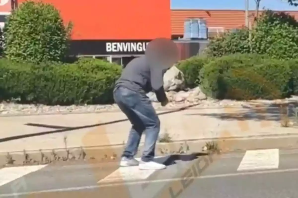Una persona amb el rostre difuminat està ajupida en una vorera a prop d'un pas de vianants, amb un fons d'arbustos i un edifici taronja que té un cartell que diu "BENVINGUTS".