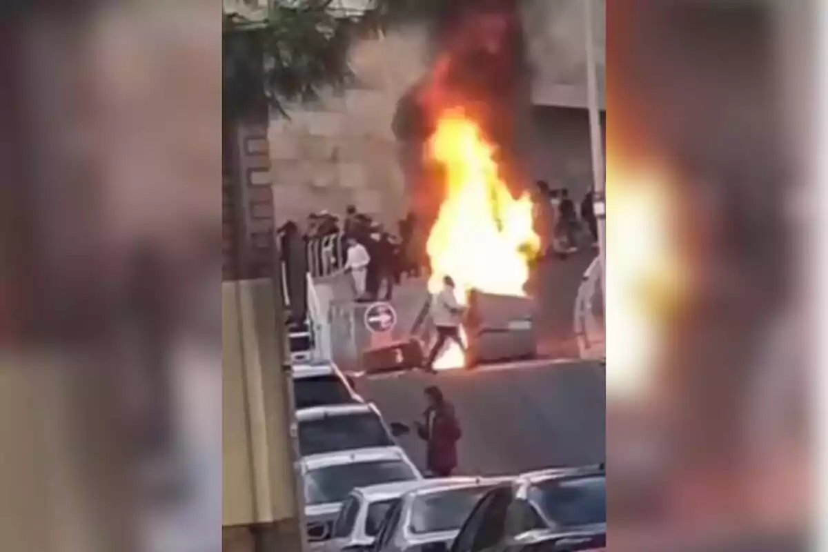 Una escena urbana muestra un contenedor en llamas rodeado de personas y vehículos estacionados.