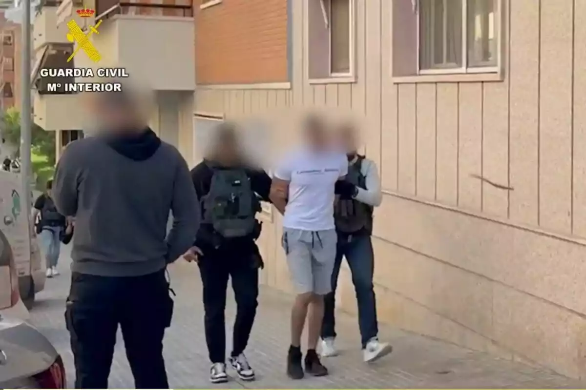 Several Civil Guard officers escort a handcuffed person along a sidewalk next to a building, with the faces of those present blurred.