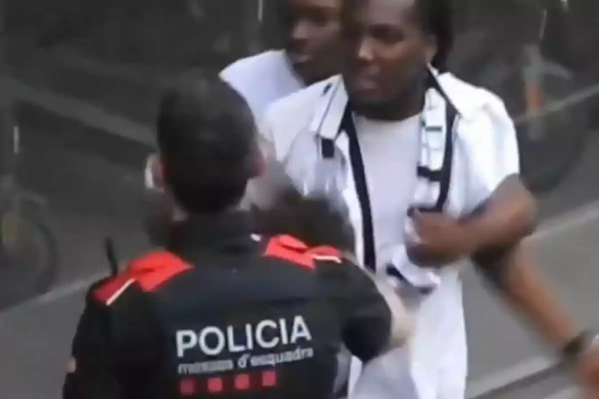 A police officer in a black and red uniform interacts with two people on the street.