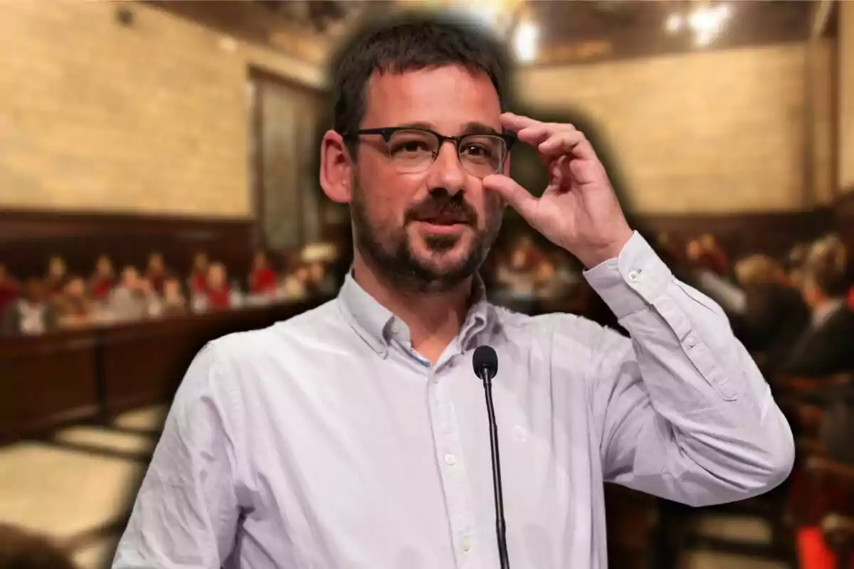 Un home amb ulleres i barba curta està dret davant un micròfon, ajustant-se les ulleres amb una mà. Porta una camisa blanca i sembla estar en una sala de reunions o conferència, amb diverses persones desenfocades al fons.