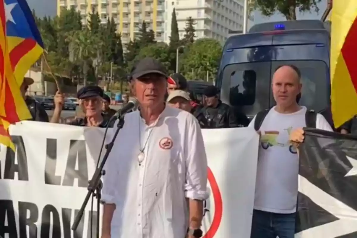 Un home amb camisa blanca i gorra negra parla davant d?un micròfon en una manifestació, envoltat de persones que sostenen banderes i pancartes.