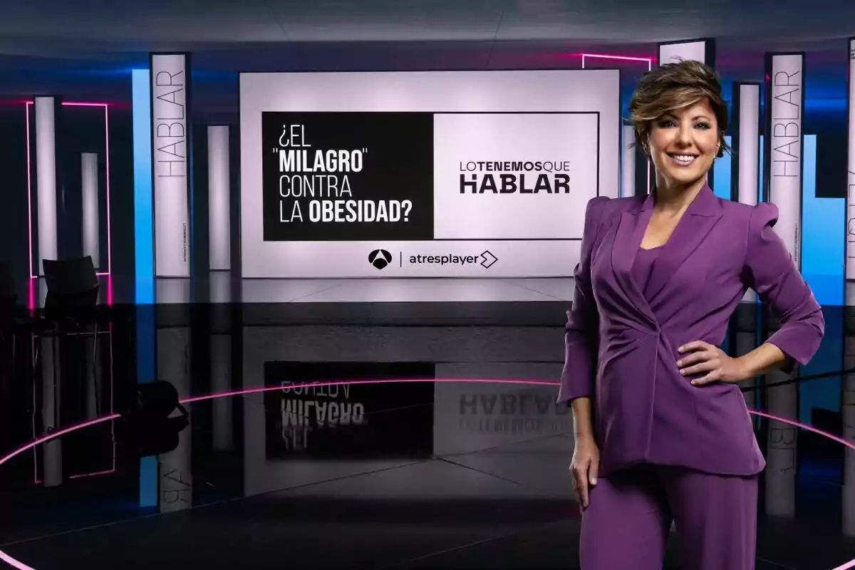 Sonsoles Ónega sonriente con un traje morado está en un set de televisión moderno con luces de neón y pantallas que muestran el texto 