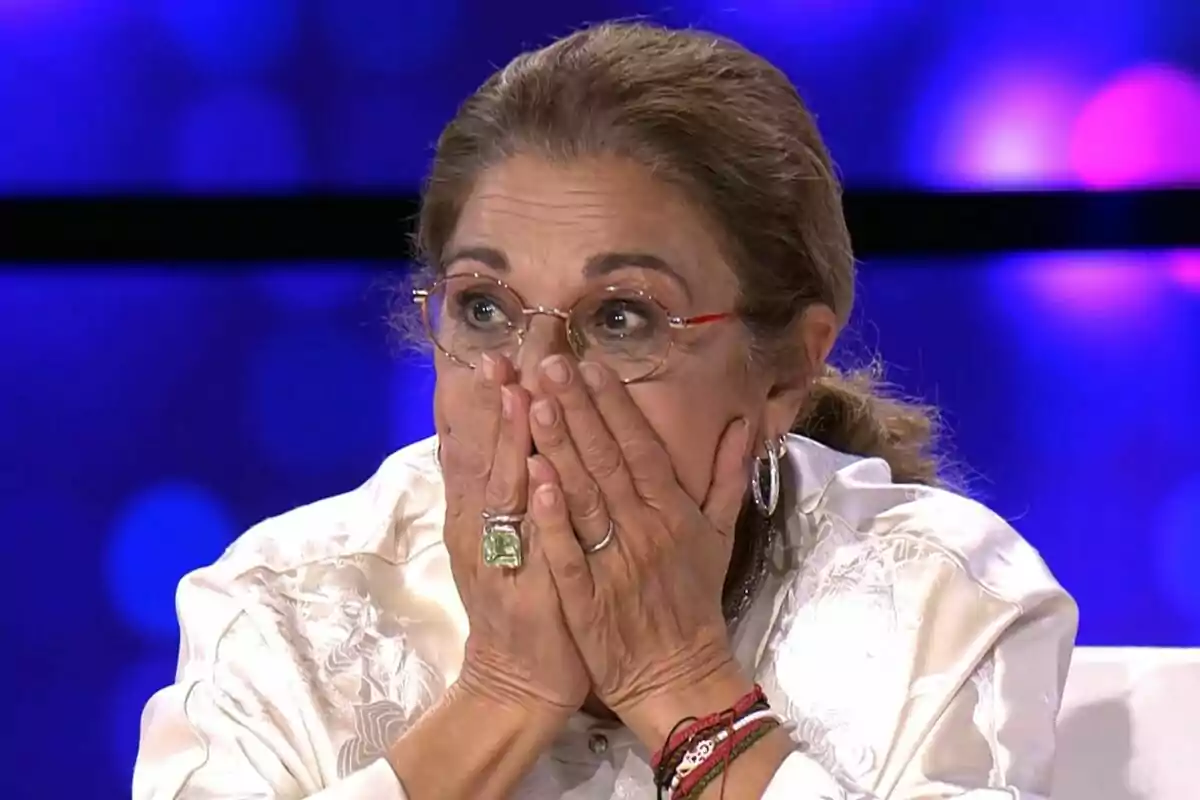 Lolita amb expressió de sorpresa es cobreix la boca amb les mans mentre està asseguda en un entorn il·luminat amb llums de fons blaves a Tu Cara Me Suena.